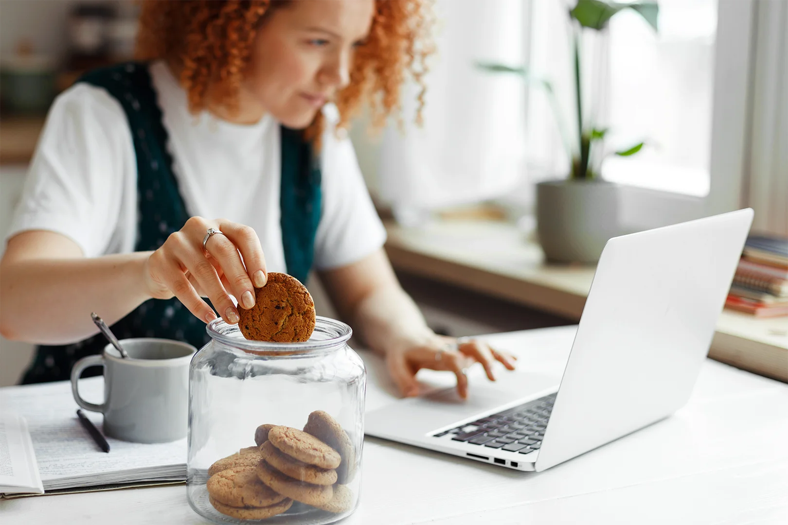 Kobieta sięgająca po ciastko z dużego słoika podczas pracy przy komputerze – spożywanie słodyczy jako czynnik ryzyka hipoglikemii reaktywnej
